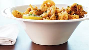 A white bowl filled with fried food, possibly calamari, garnished with herbs and lemon slices, placed on a wooden table.