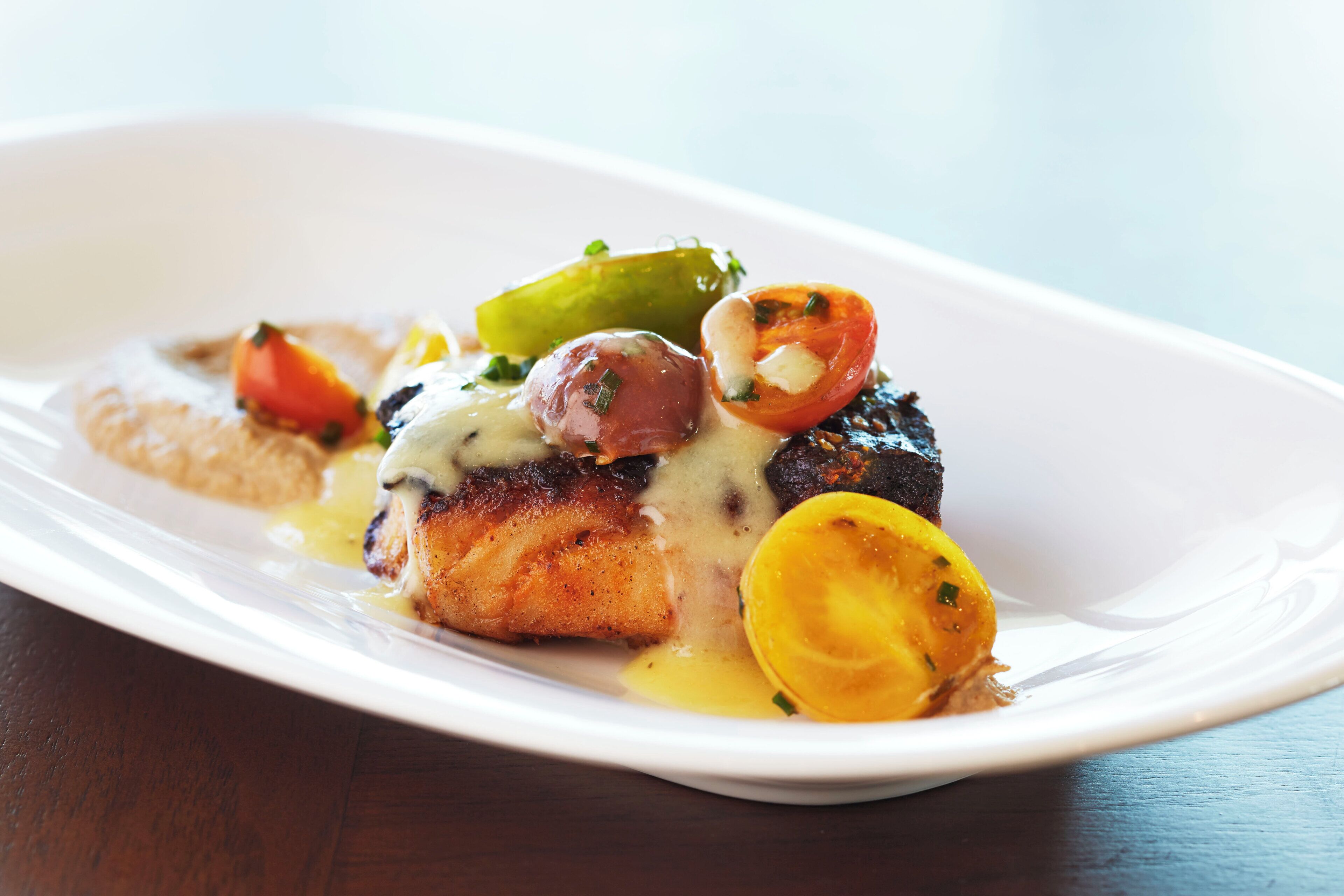 A white plate with a piece of grilled fish topped with a creamy sauce, garnished with cherry tomatoes and a slice of lemon.