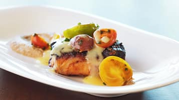A white plate with a piece of grilled fish topped with a creamy sauce, garnished with cherry tomatoes and a slice of lemon.