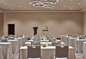 Sala de conferência com mesas preparadas para uma reunião, um púlpito e um vaso decorativo com plantas.