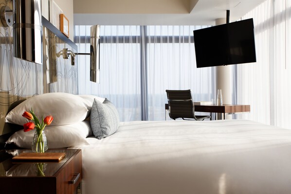 A hotel room with a large bed, a desk with a chair, a flat-screen TV, and a vase with flowers on the nightstand.