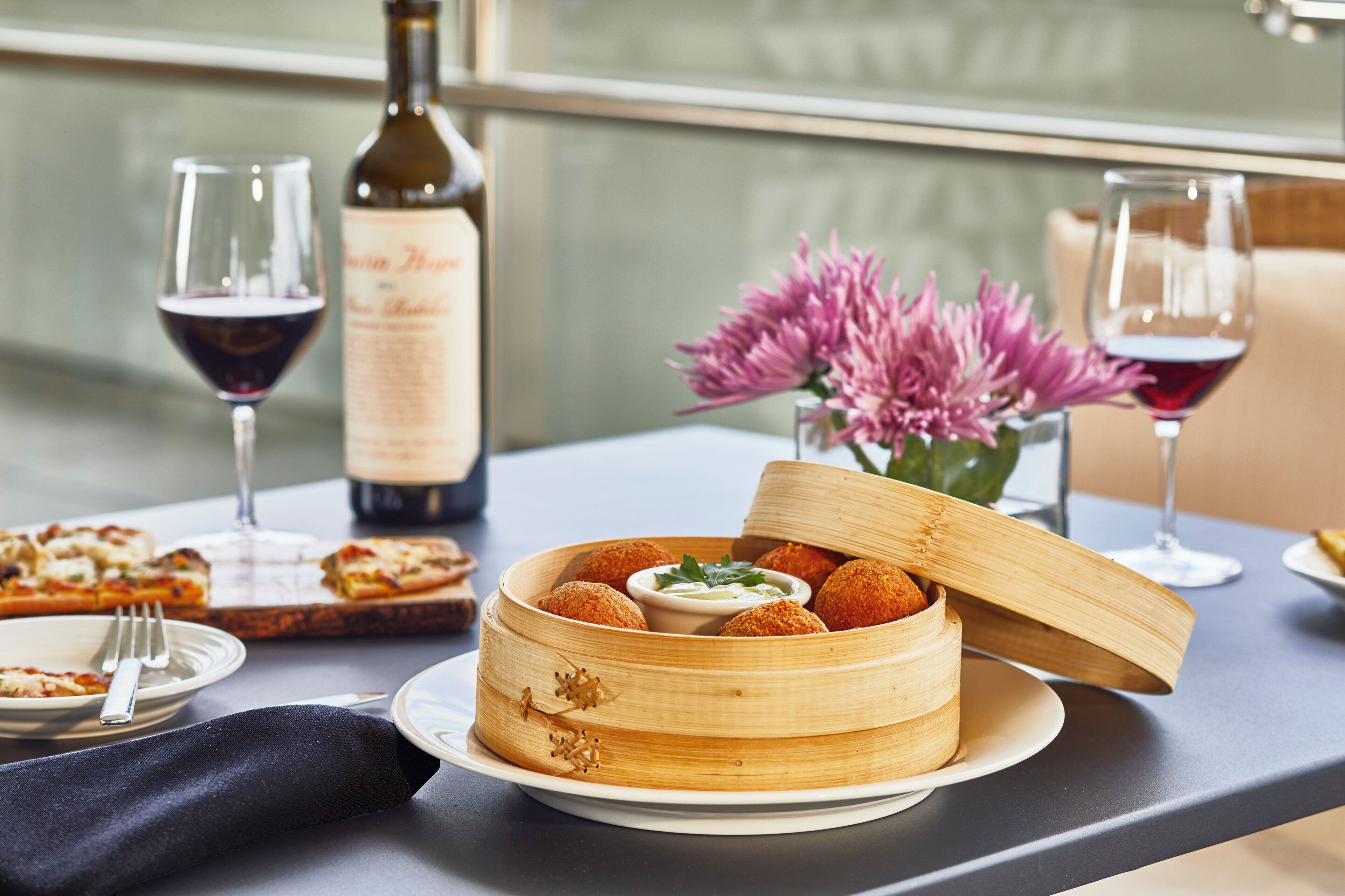 A dining table with a wooden basket containing food items, a bottle of wine, and glasses of red wine.