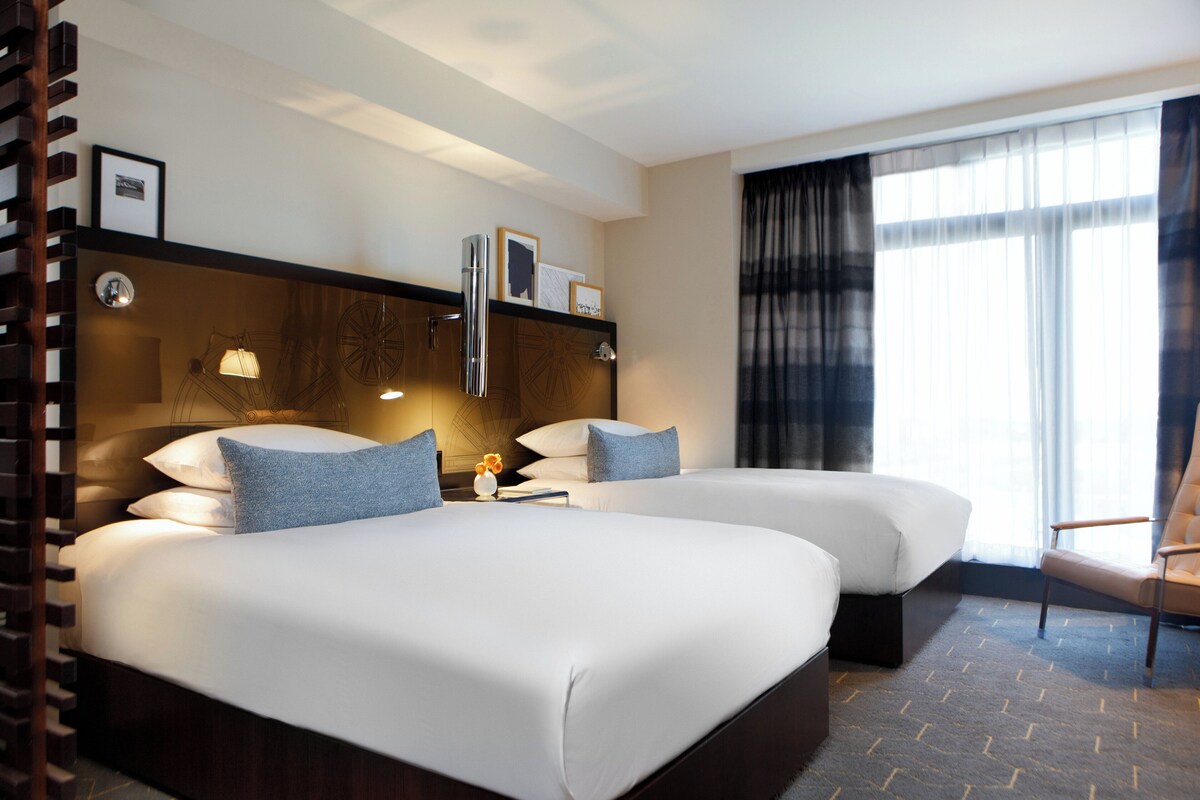 Habitación de hotel con dos camas, un ventanal con cortinas, cabecero de madera y una repisa con objetos.