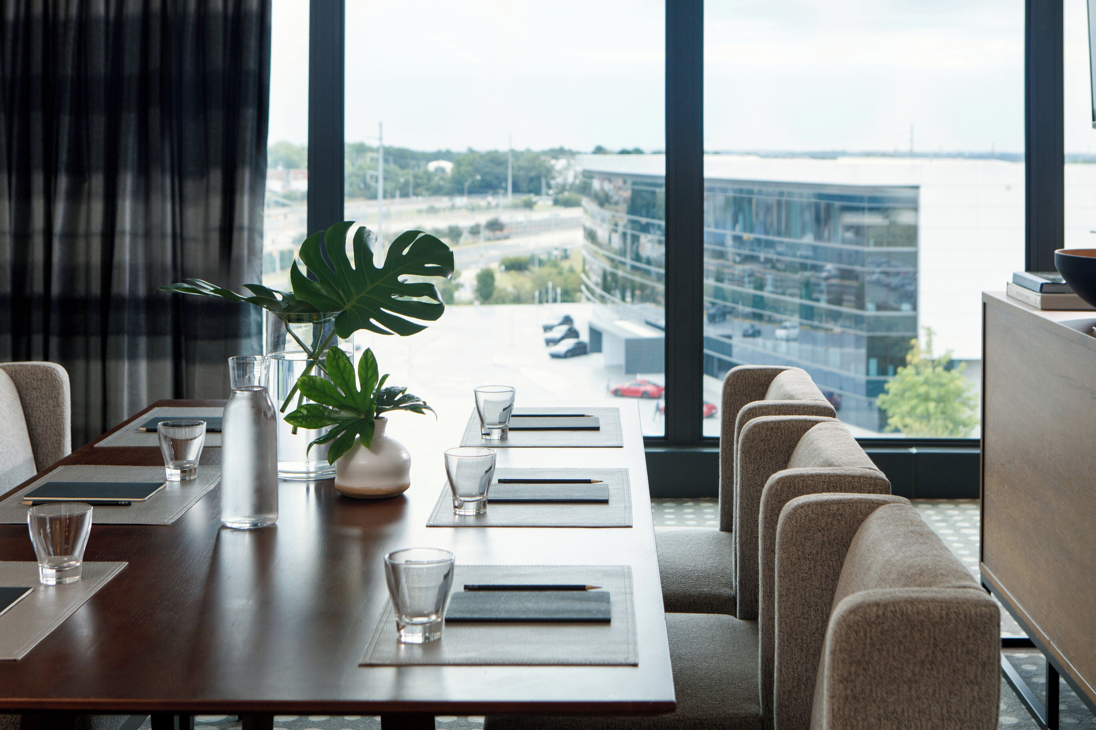 A modern hotel dining area with a long table set for a meal, a potted plant, and a view of a cityscape through large windows.
