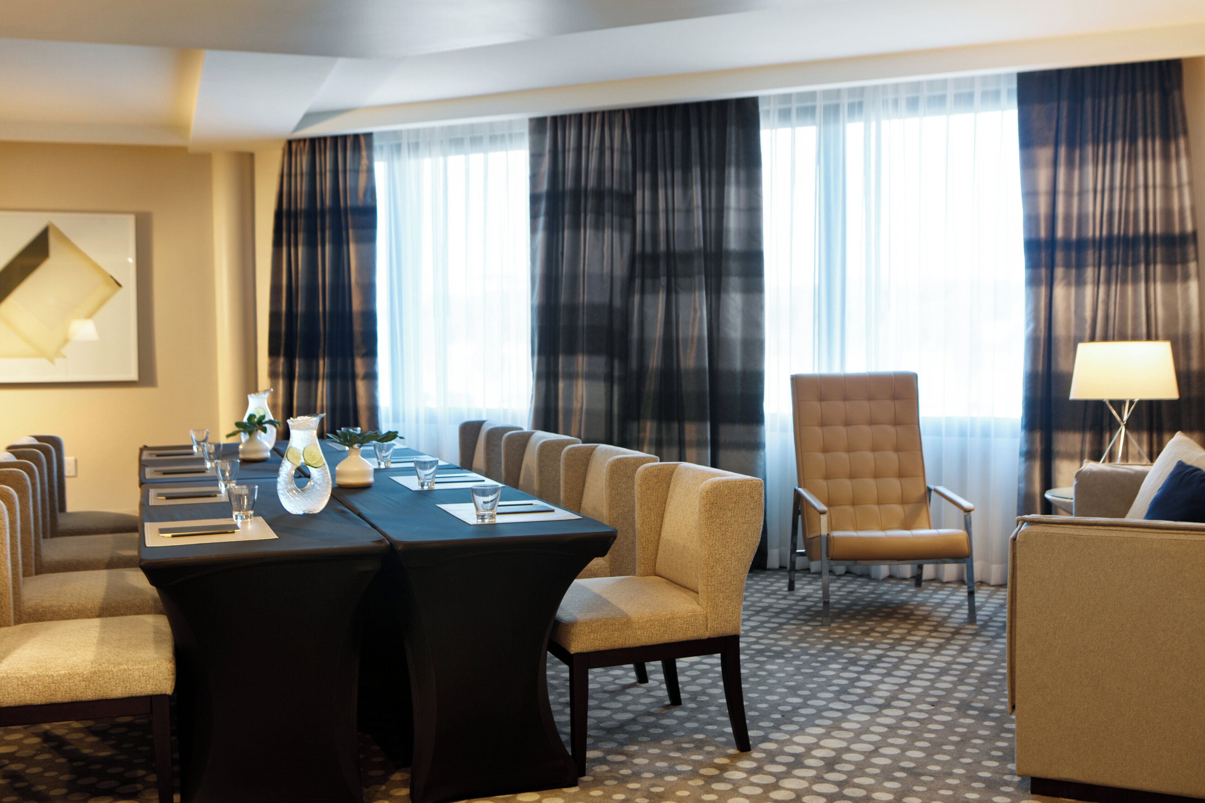 A hotel room with a conference table, chairs, a sofa, a lounge chair, and a lamp.