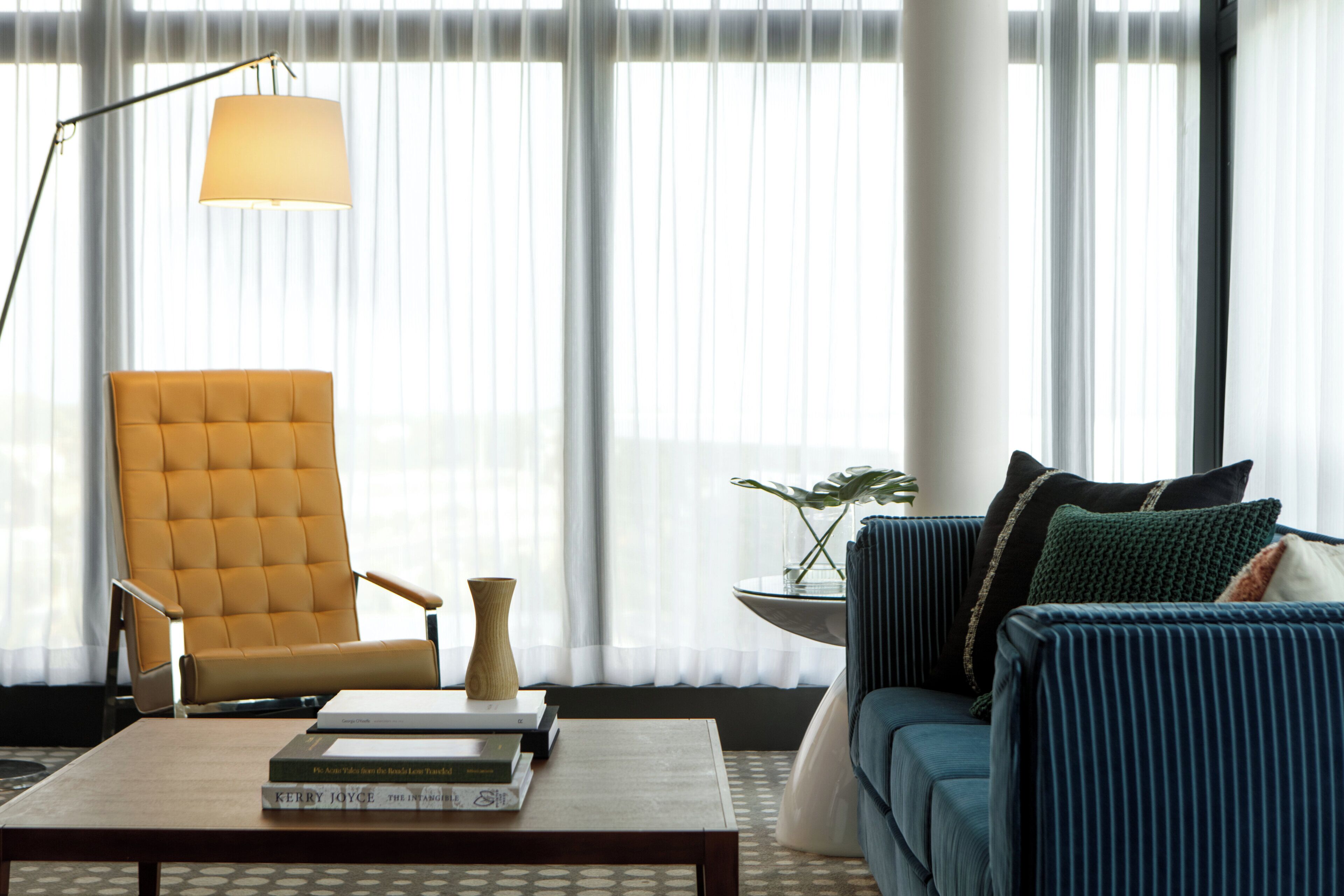 A modern living room with a yellow armchair, a blue sofa, a coffee table with books, and a floor lamp.