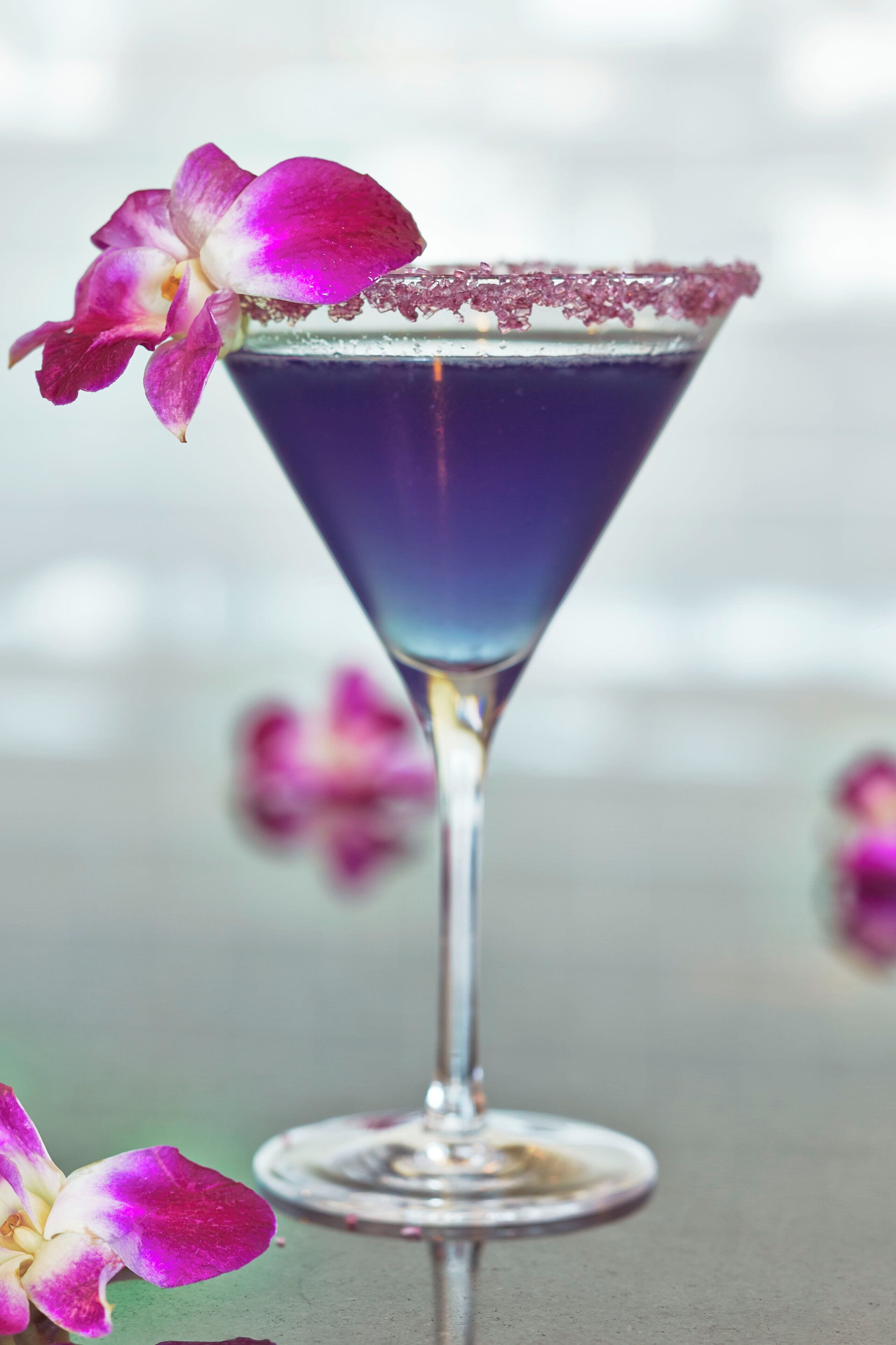 Een martini glas met een paars drankje, een roze bloem op de rand, en een onscherpe achtergrond met meer bloemen.