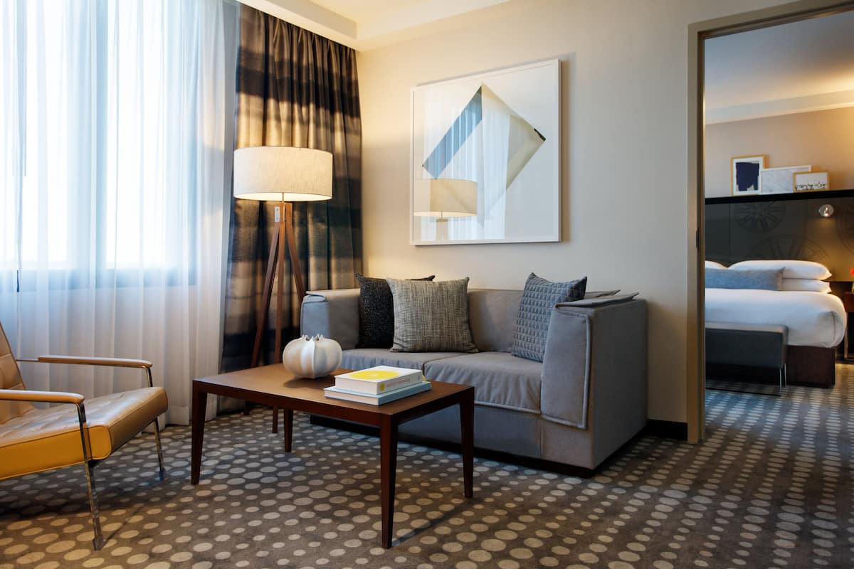 A hotel room with a sofa, a coffee table with a book, a lamp, and a large window with curtains.