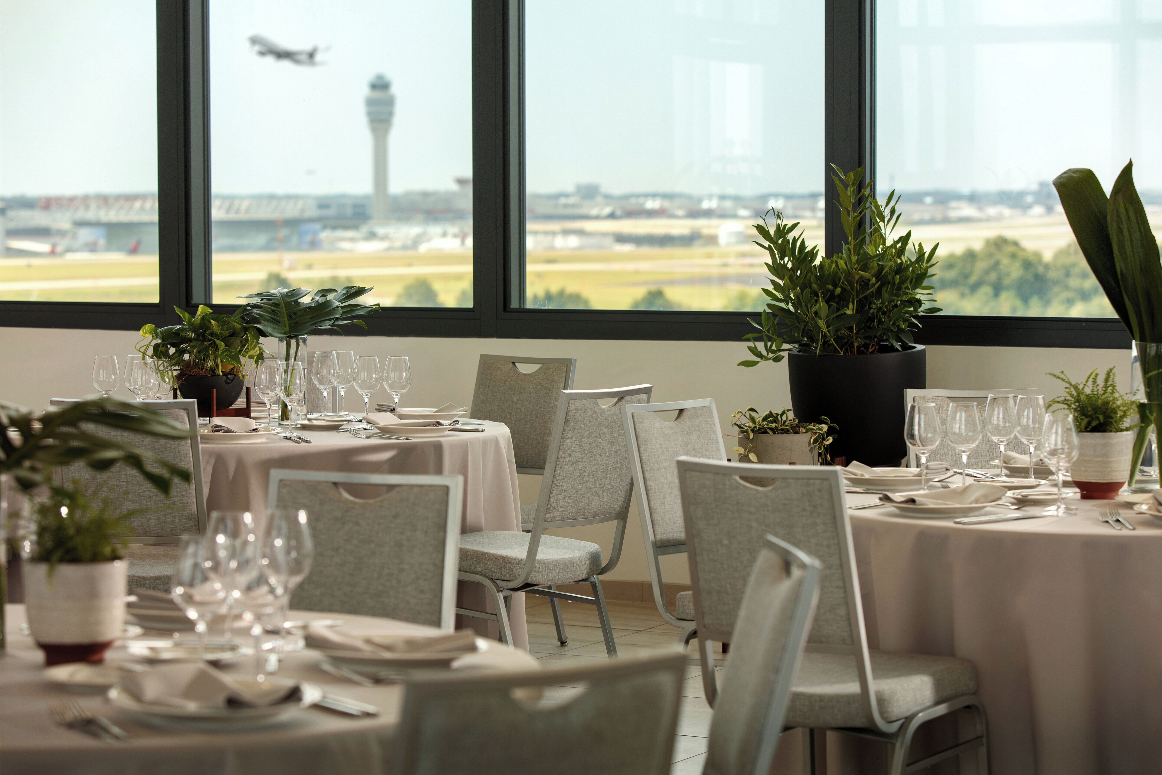 A dining area with tables set for a meal, potted plants, and a view of an airport in the background.