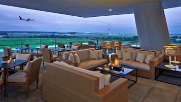 A rooftop lounge with wicker furniture, a fire feature, and a view of an airport.