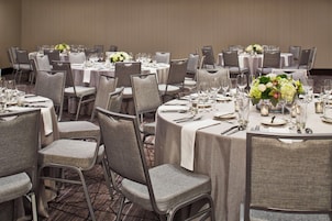 Un salón de banquetes con mesas redondas dispuestas para el servicio de comidas, cada una con mantel blanco, sillas y un centro de mesa.