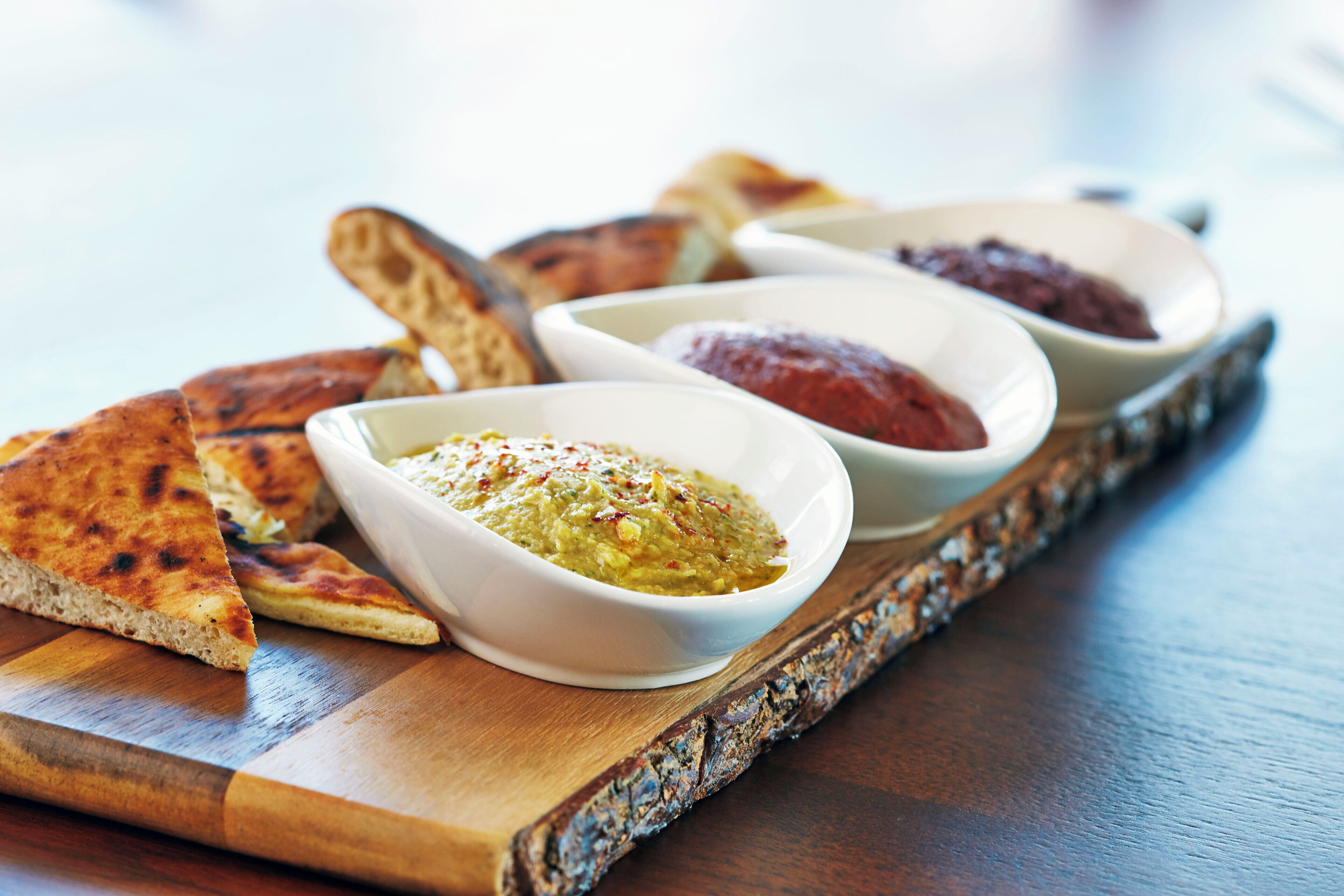 A wooden serving board with flatbreads and three small bowls of dips.