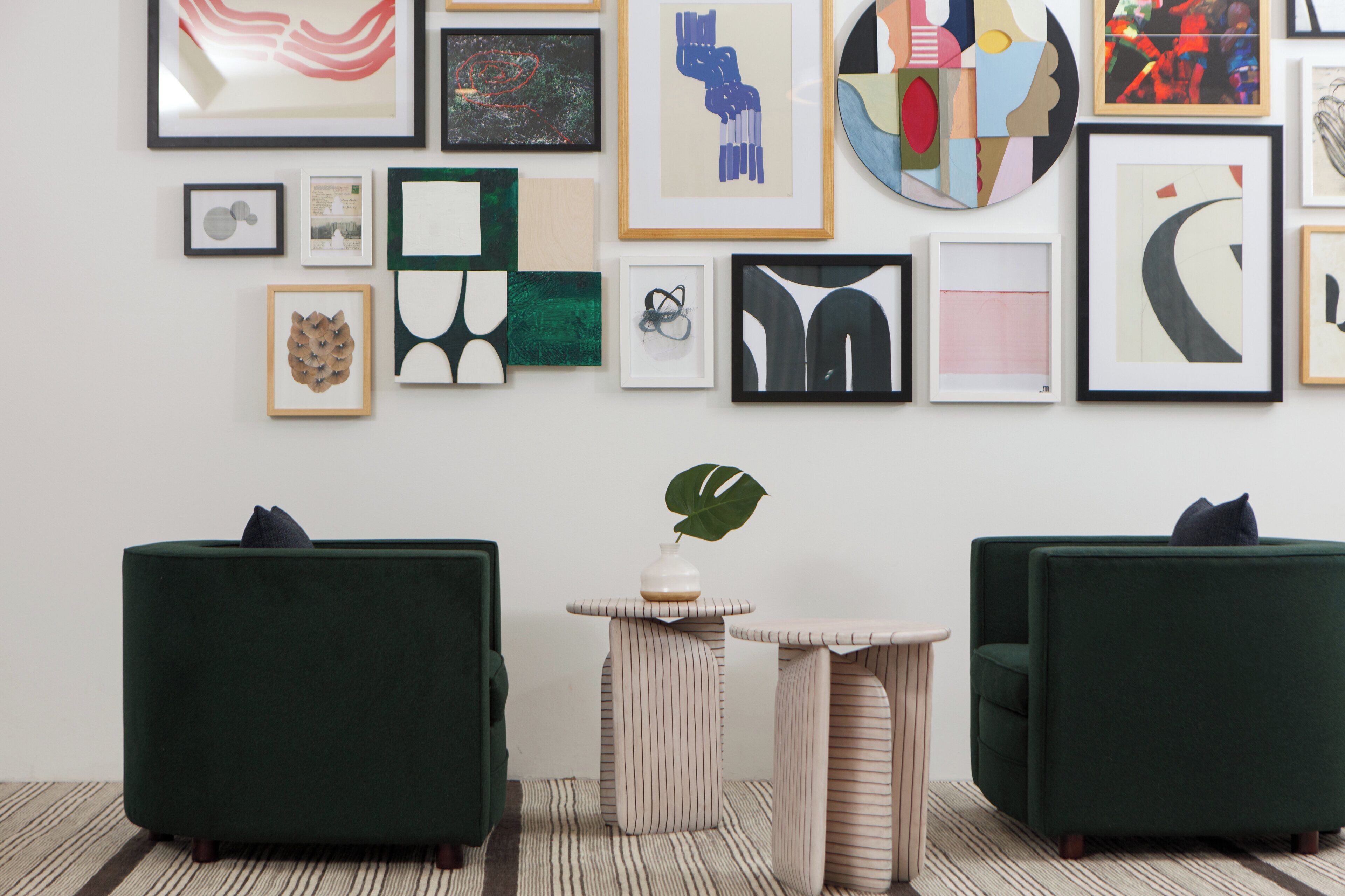 A room with two green armchairs, a small round table with a plant, and a wall adorned with various framed artworks.