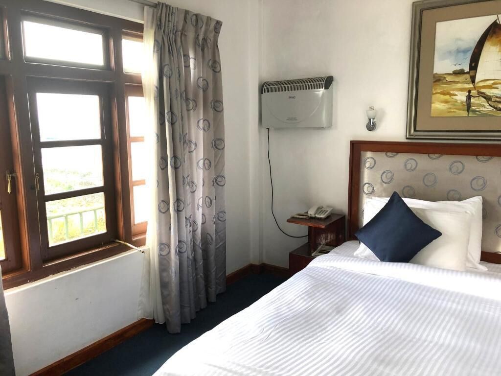 Een hotelkamer met een bed, een raam met gordijnen, een airconditioningapparaat, een nachtkastje met een telefoon en een schilderij aan de muur.