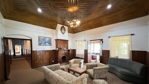 Un salón espacioso con chimenea, paneles de madera y una araña. Hay varias zonas de estar con sofás estampados y un ventanal con cortinas.