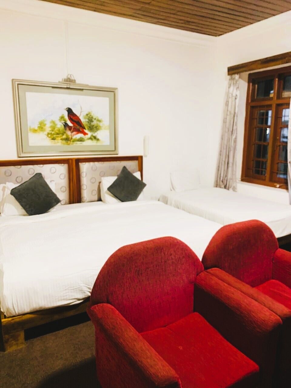 A room with two beds, a red chair, a framed picture on the wall, and a window with curtains.