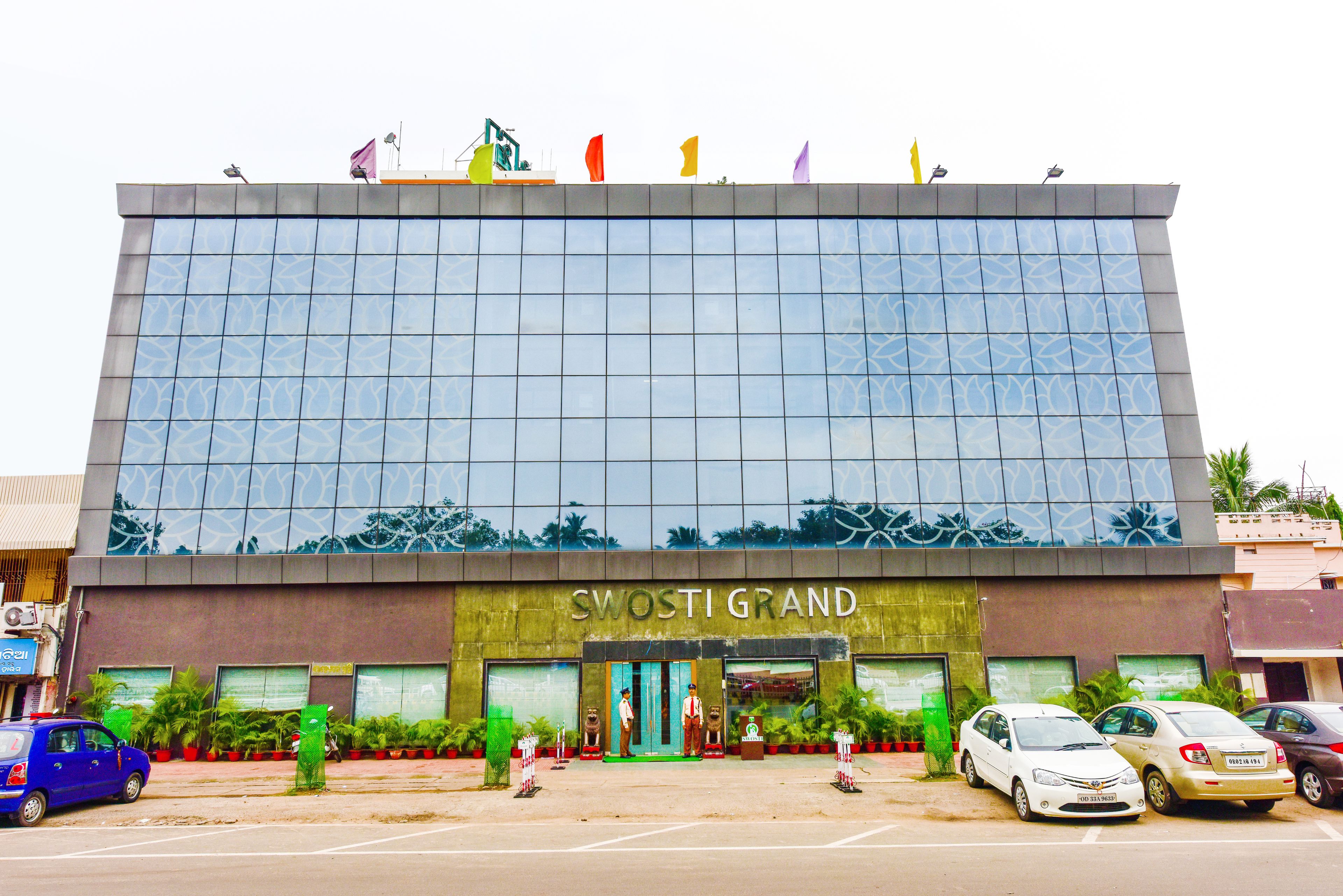Un edificio moderno con fachada de vidrio reflectante y el nombre “SWOSTI GRAND” en la planta baja. Varias automóviles están estacionadas frente al edificio, y plantas en macetas decoran la entrada.