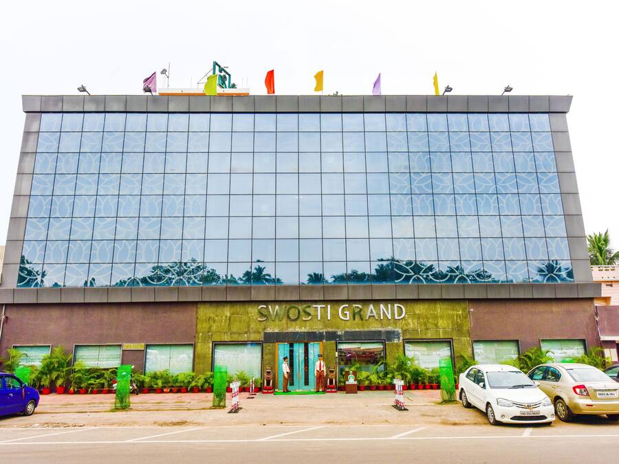 A modern building with a reflective glass facade and the name "SWOSTI GRAND" on the ground floor. There are several cars parked in front, and potted plants are arranged along the entrance.