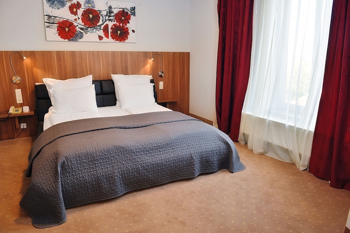 A neatly made bed with a grey quilt, a wooden headboard, and a wall-mounted artwork of red flowers.