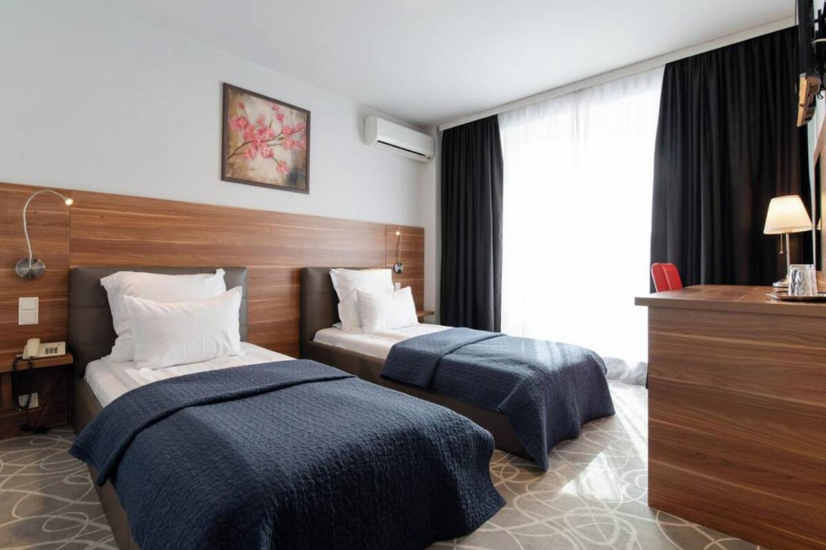 A hotel room with two beds, a desk, a lamp, and a painting on the wall.
