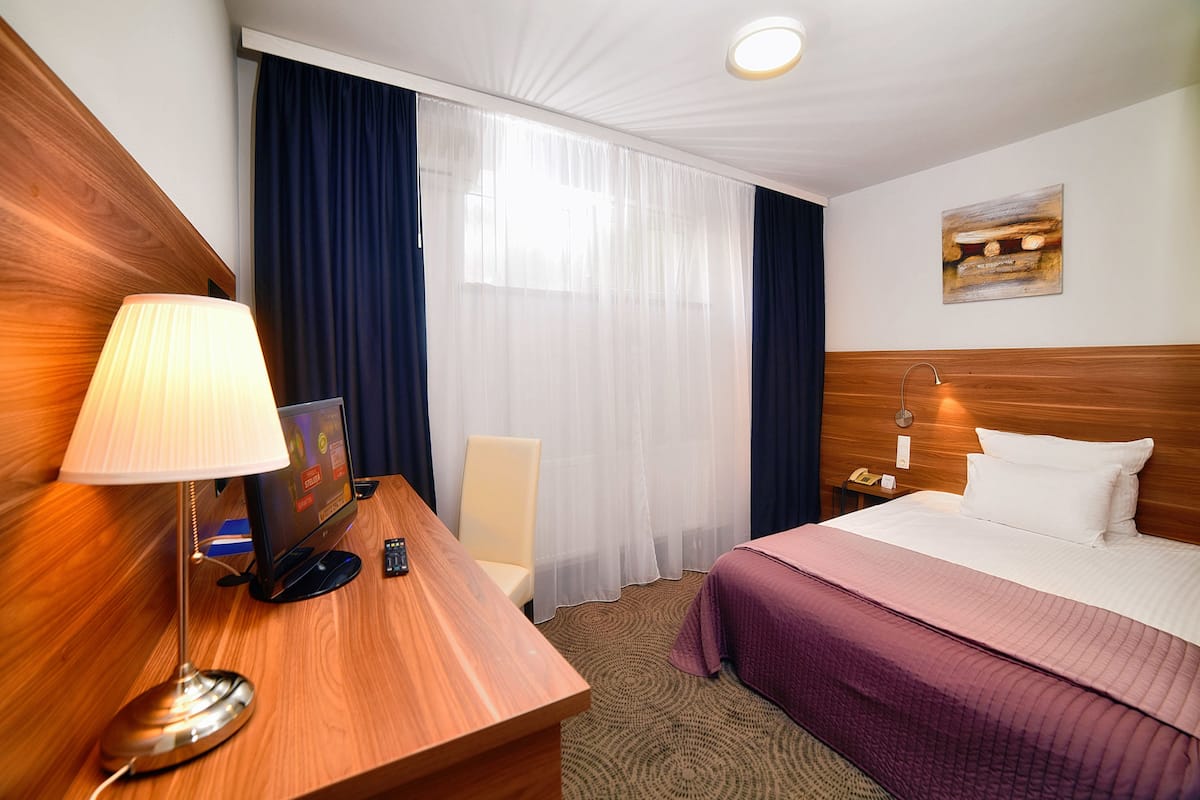 A hotel room with a wooden desk, a lamp, a bed with a purple bedspread, a chair, a nightstand with a phone, and a painting on the wall.