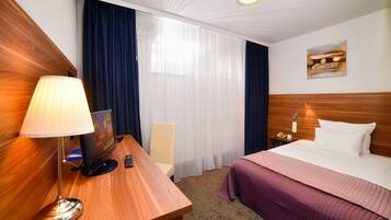 A hotel room with a wooden desk, a lamp, a bed with a purple bedspread, a chair, a nightstand with a phone, and a painting on the wall.