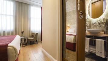 A hotel room with a bed, a chair, a small table with a vase, and a bathroom with a sink and mirror.