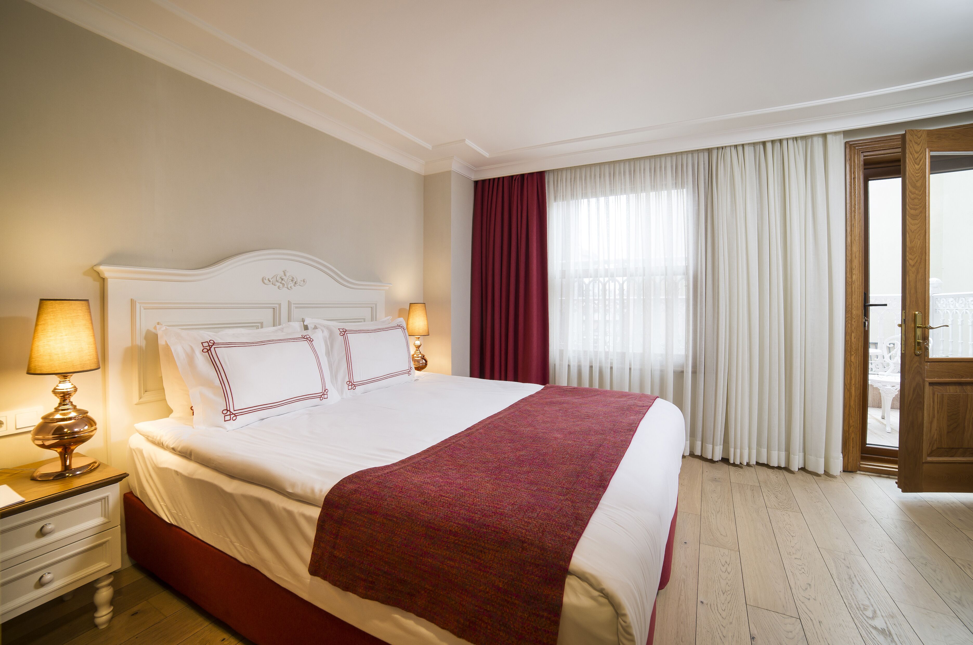 Una cama bien hecha con sábanas blancas y una manta roja, una mesita de noche de madera con una lámpara, y vistas a un balcón a través de una ventana con cortinas rojas.