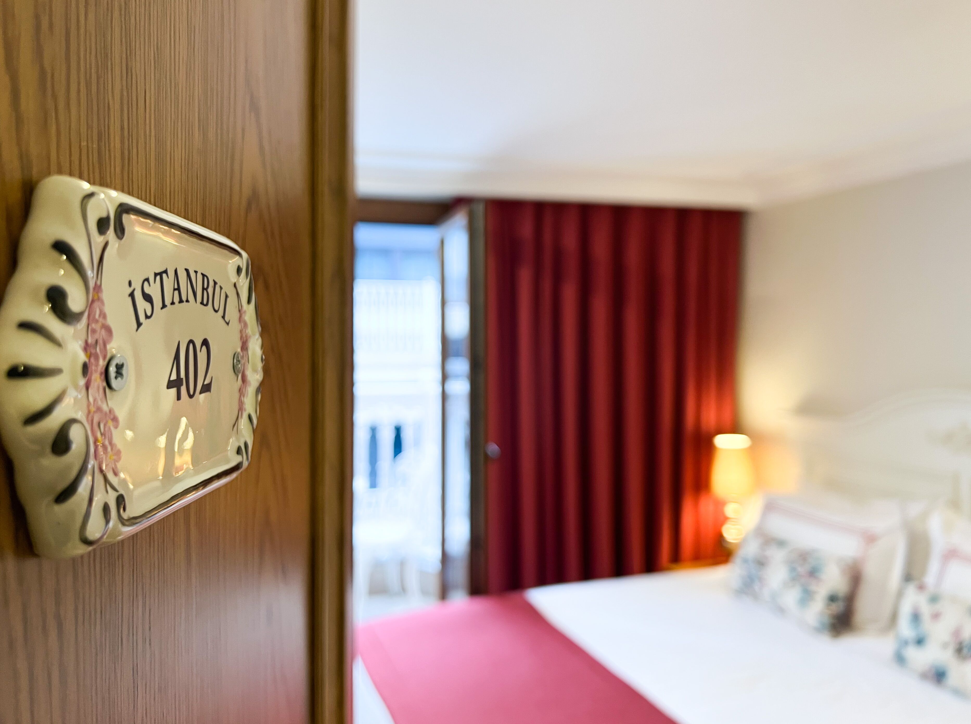 A hotel room with a bed, a red curtain, and a door with a sign reading "ISTANBUL 402".