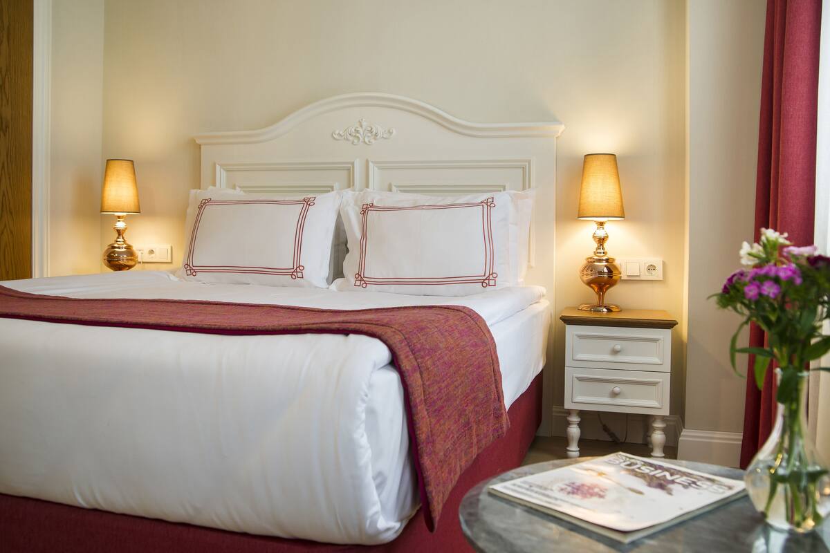 A neatly made bed with white linens and a red blanket, two bedside lamps, a small bedside table with a magazine, and a vase with flowers.