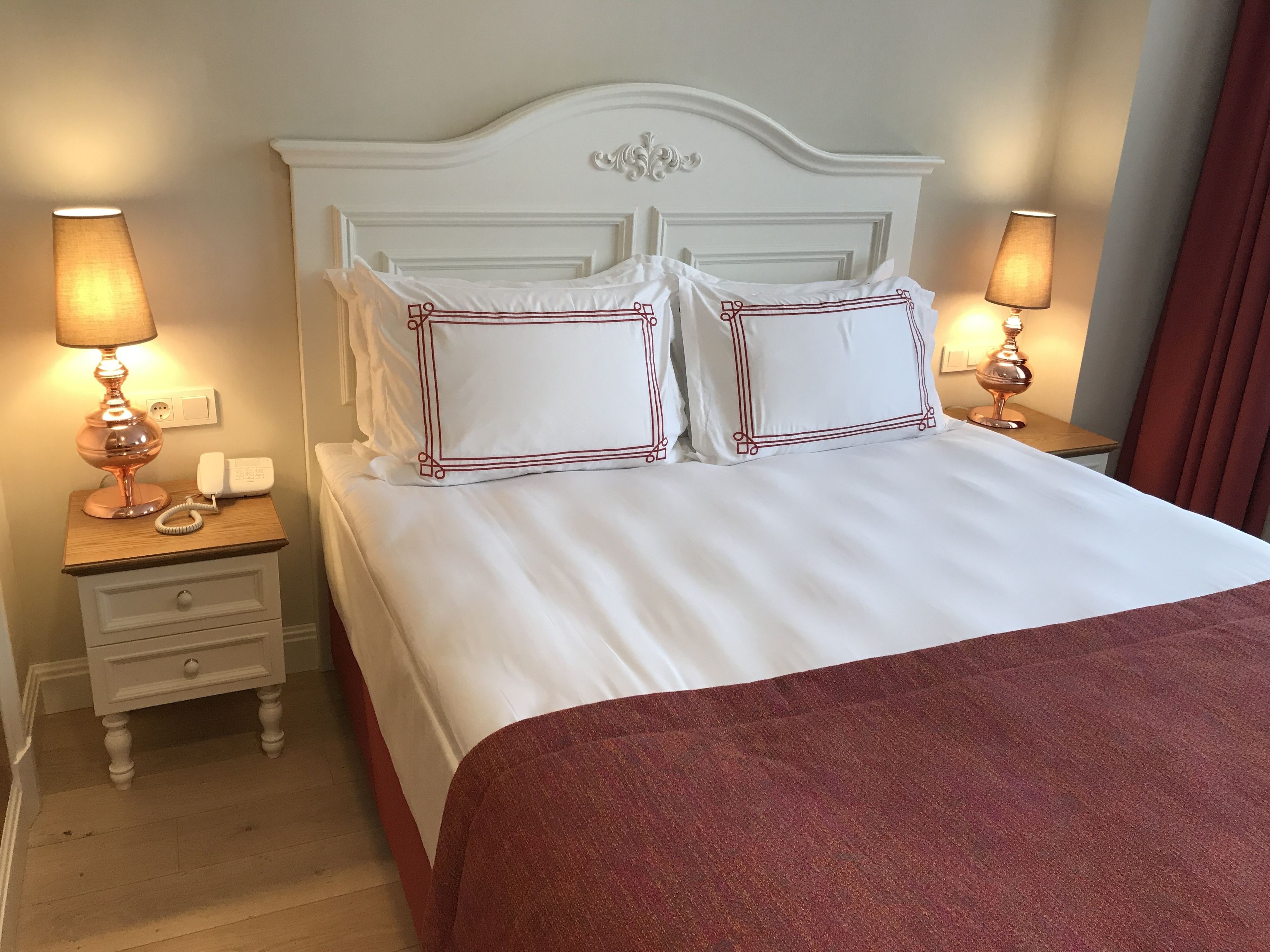 Cama bem arrumada com lençóis brancos e almofadas com detalhes vermelhos, acompanhada por duas mesas de cabeceira com abajures, uma das quais possui um telefone.