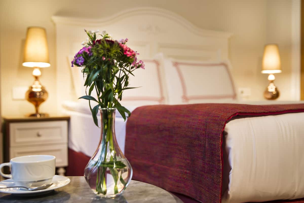 A hotel room with a bed, a bedside table with a lamp, a vase with flowers, and a cup on the table.