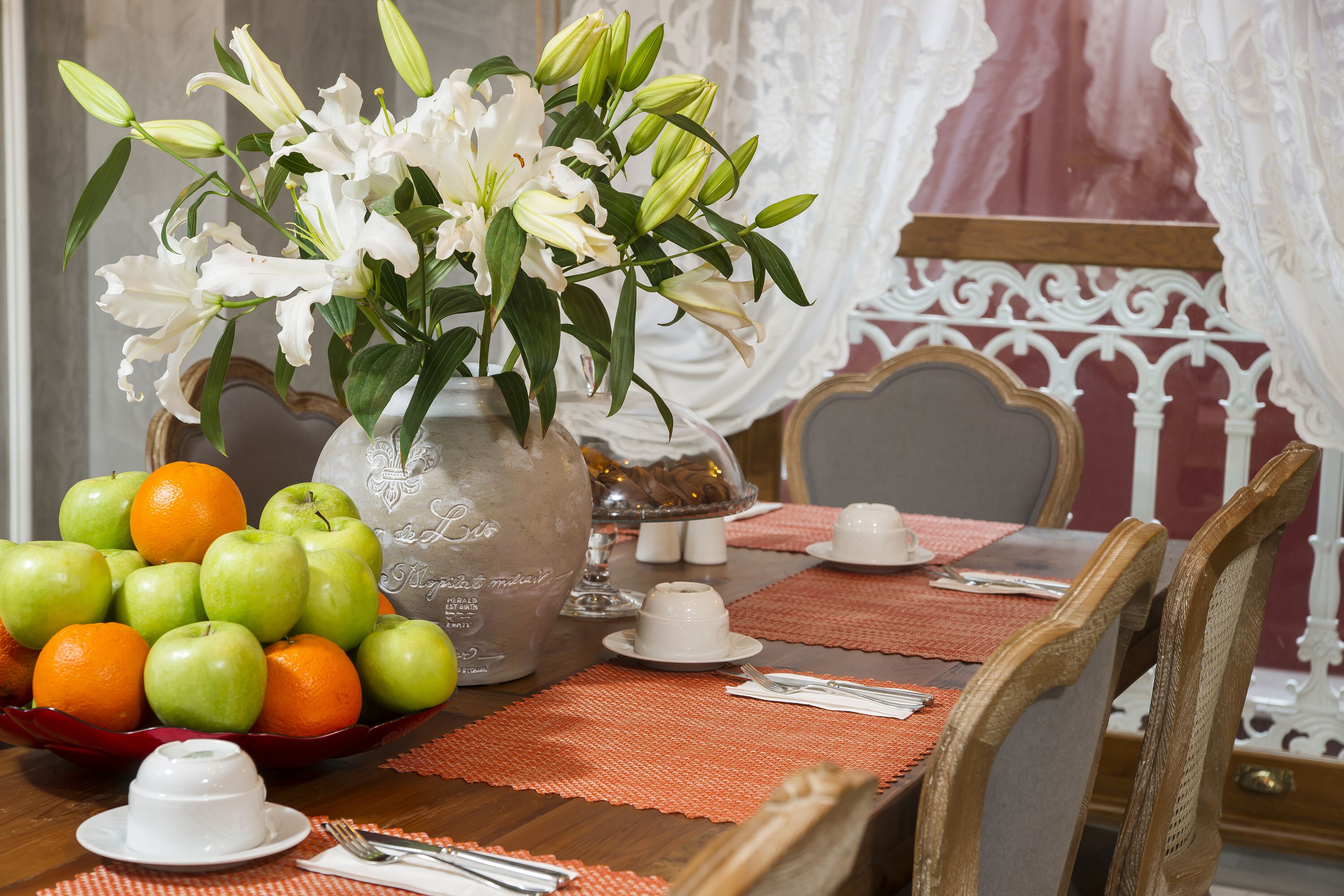 A dining table with a vase of white lilies, a bowl of green apples and oranges, and a clear glass pitcher.