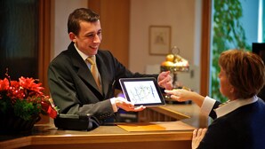 Un comptoir d’accueil où un homme tient une tablette affichant une carte, une femme la reçoit, et une plante en pot avec des fleurs rouges se trouve à gauche.
