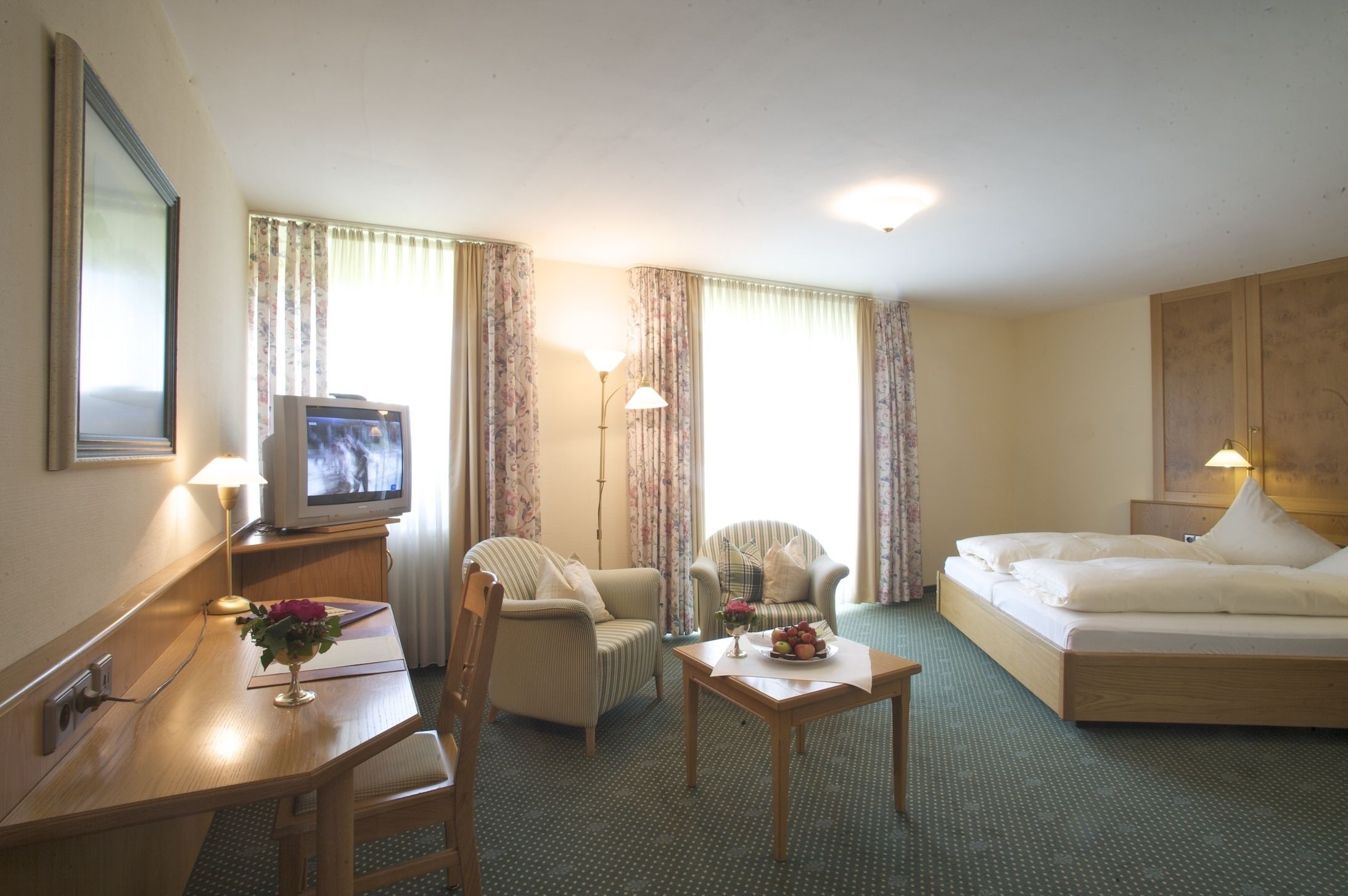 A hotel room with a bed, two armchairs, a desk with a television, and a small table with a vase of flowers.