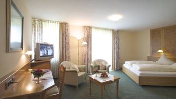 Una habitación de hotel con una cama, dos sillones, un escritorio con televisor y una mesita con un jarrón de flores.