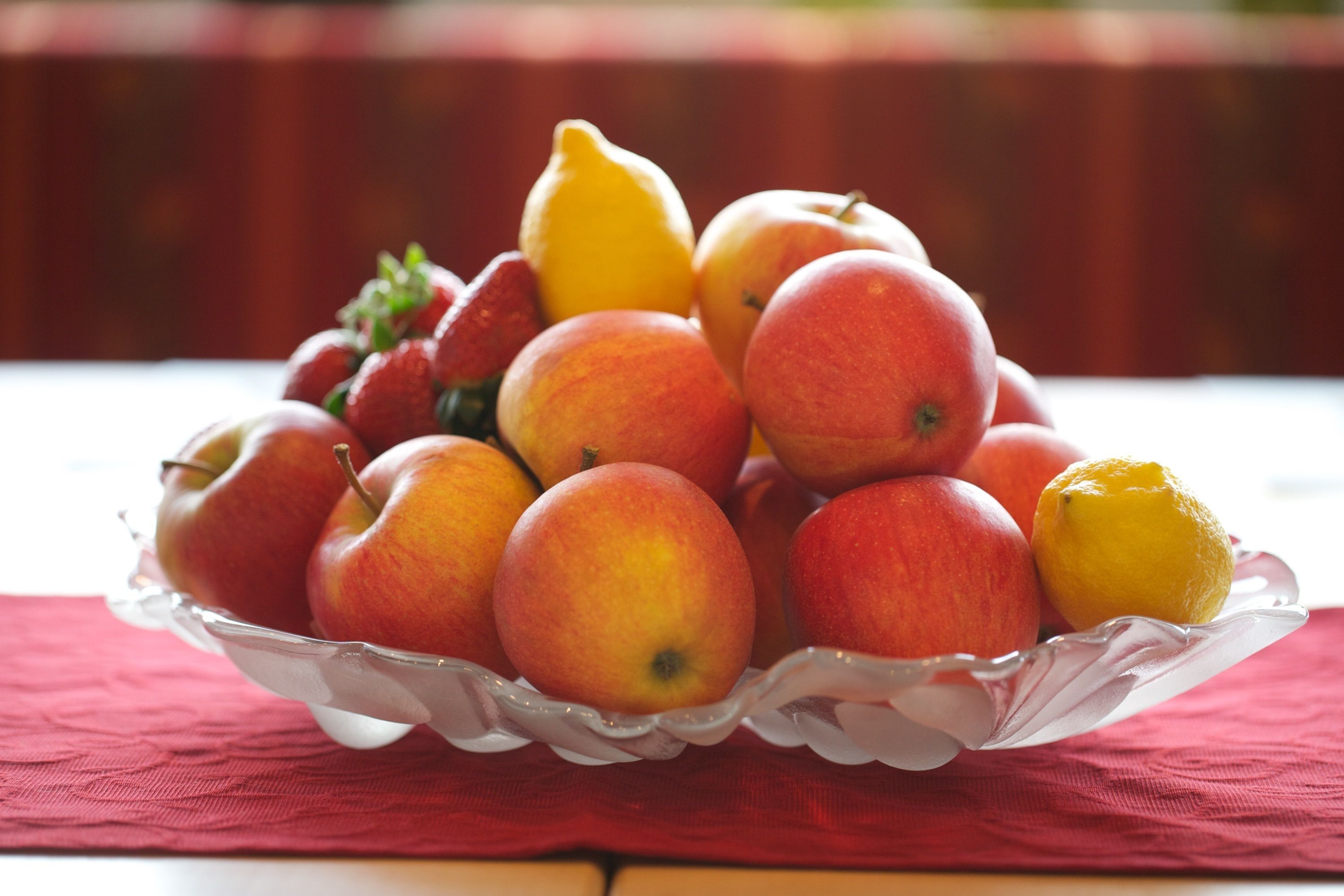 A glass bowl filled with apples and a lemon on a red cloth.