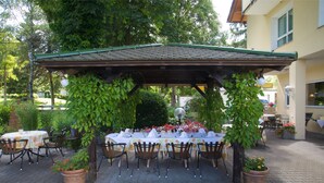 Une terrasse extérieure avec des tables et des chaises sous une pergola en bois, entourée de plantes en pot et dotée d’un sol en briques.