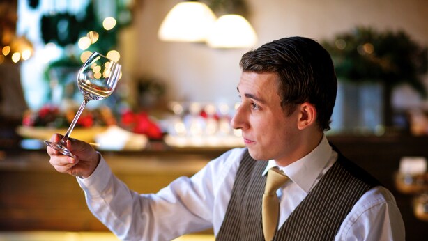 Une personne tenant un verre de vin, portant un badge nominatif, dans un décor intérieur chaleureux et bien éclairé, avec des éléments d'arrière-plan flous.