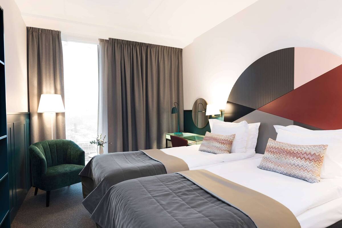 Quarto de hotel com duas camas, uma poltrona verde, uma mesa de trabalho com luminária e uma janela ampla com cortinas.
