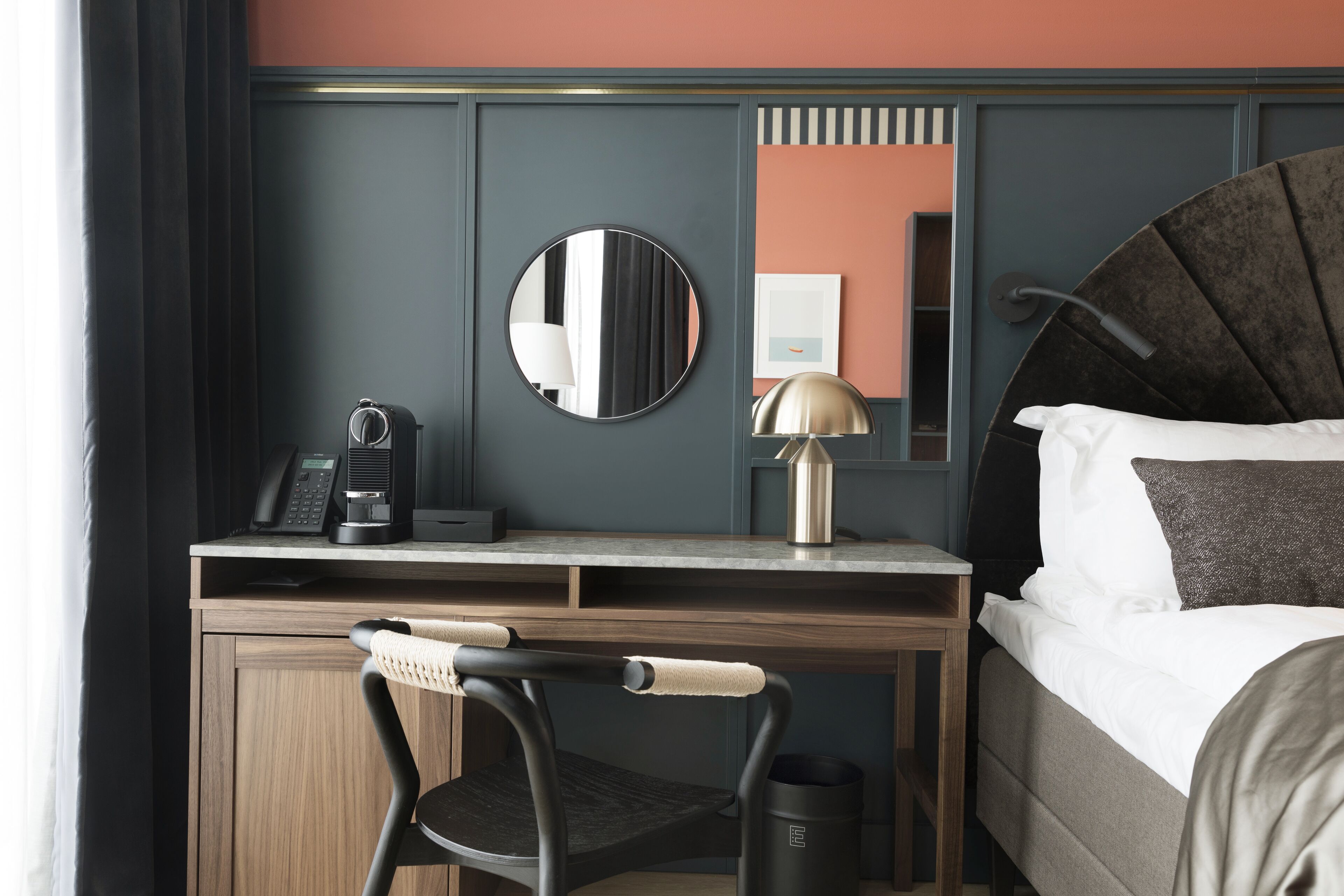 Habitación de hotel con escritorio de madera, una silla, cama con ropa de cama blanca y gris, un espejo redondo, lámpara y cafetera.
