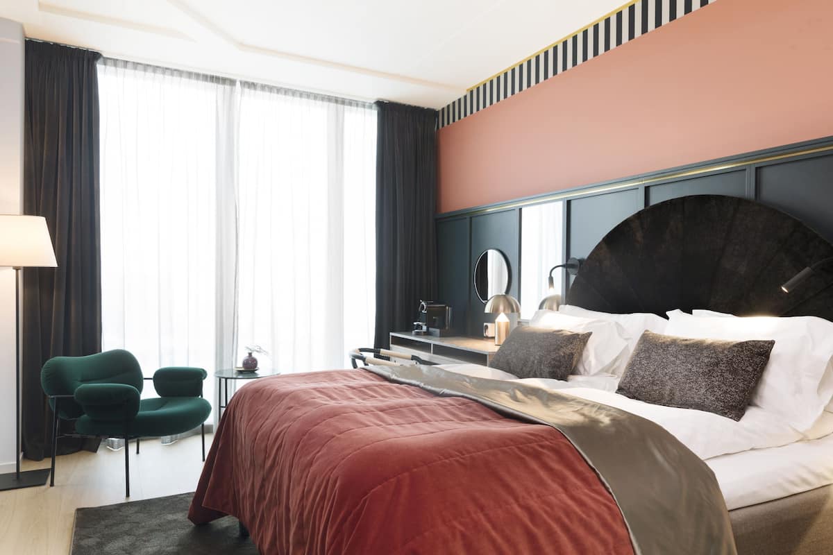 Quarto de hotel com uma cama grande, uma escrivaninha, uma cadeira e uma janela ampla com cortinas.