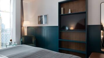 A hotel room with a bed, a chair, a desk, a bookshelf, and a window with a view of buildings.