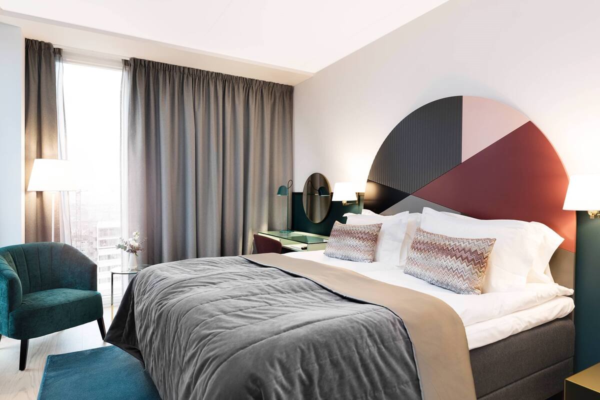 Quarto de hotel moderno com uma cama grande, uma escrivaninha, uma cadeira e vista para a cidade.