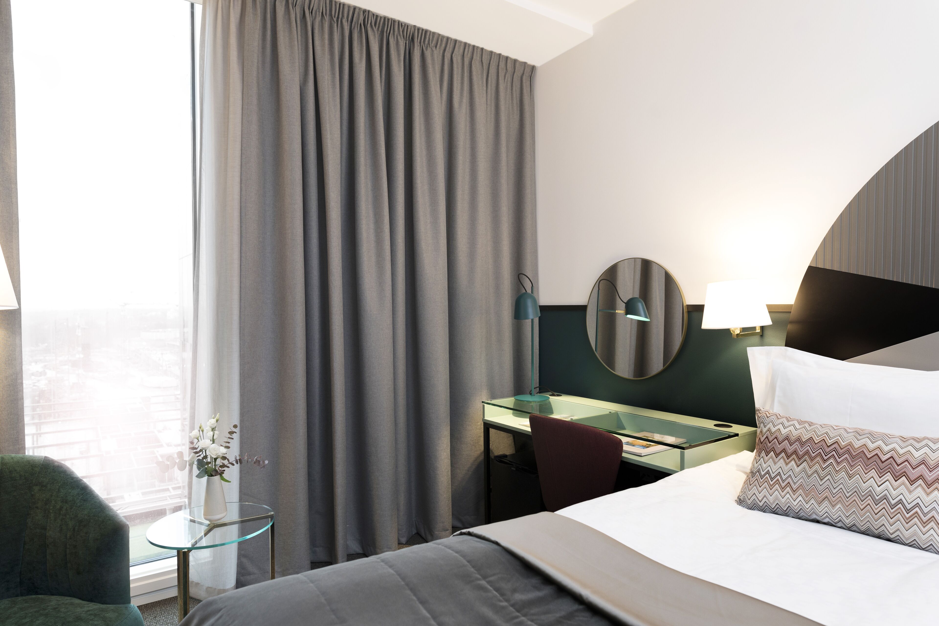 A hotel room with a bed, a desk, a chair, a glass table, a mirror, and a view of the city.
