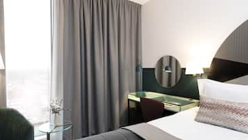A hotel room with a bed, a desk, a chair, a glass table, a mirror, and a view of the city.