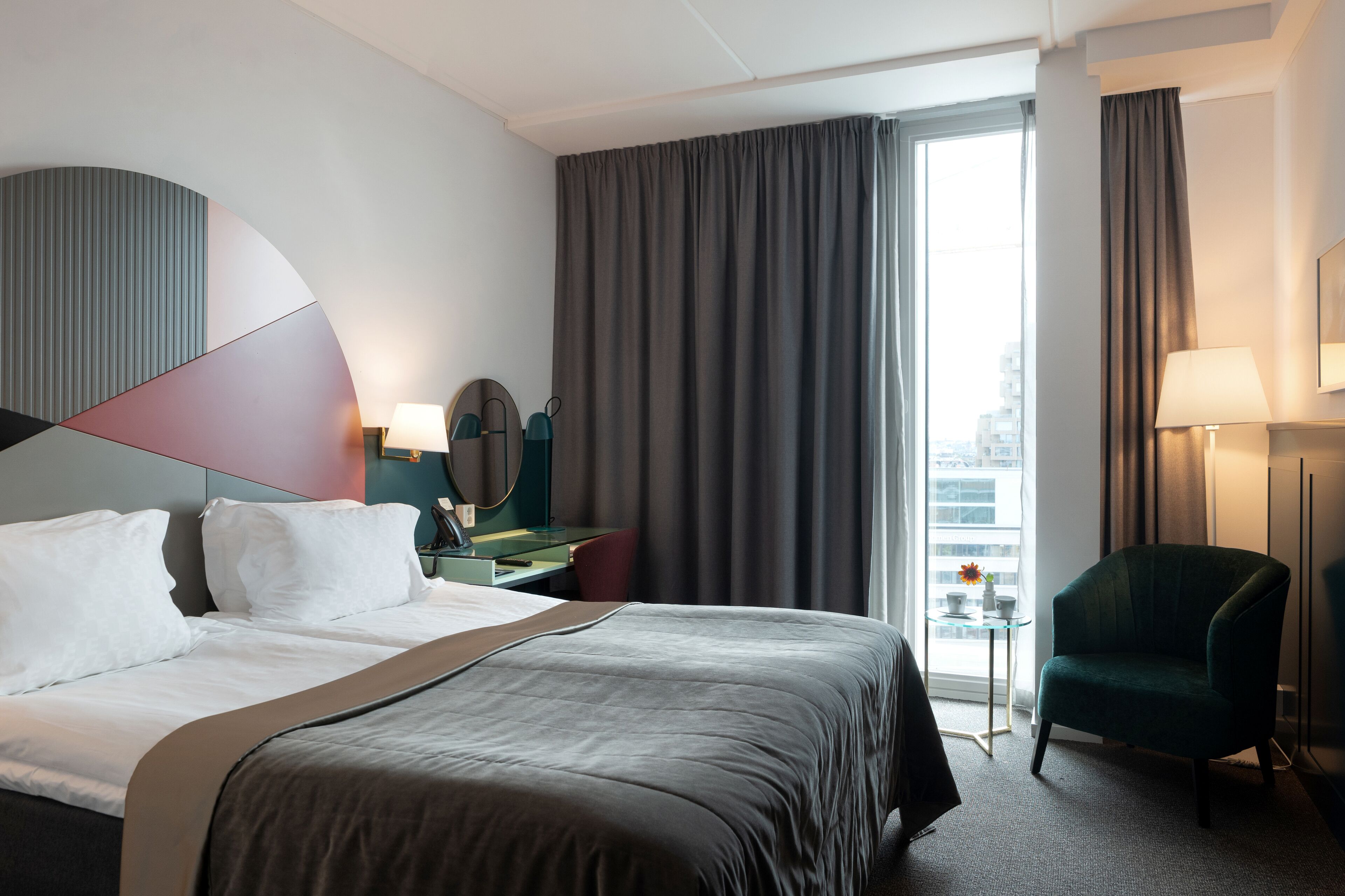 A hotel room with a large bed, a desk with a lamp, a chair, and a view of the city through the window.