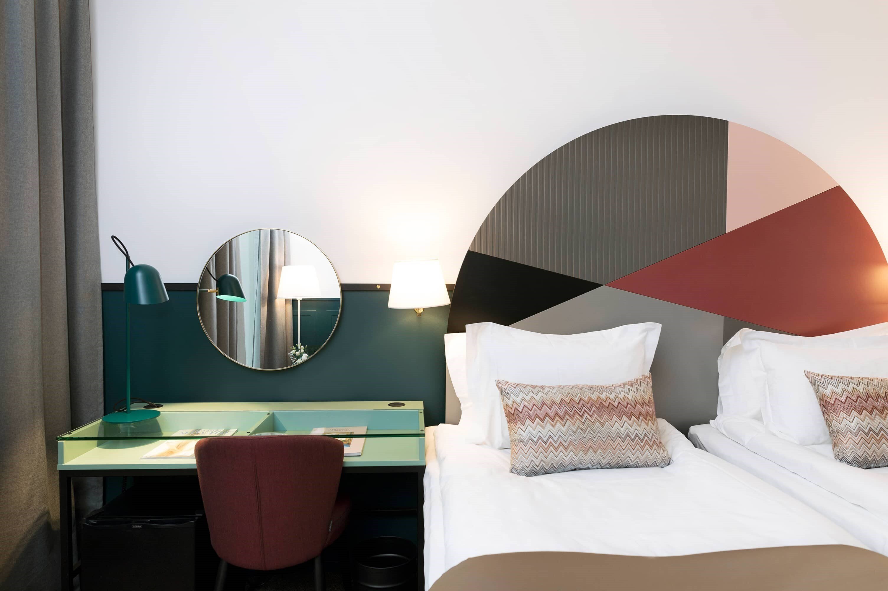 A hotel room with a bed, a desk, a chair, a mirror, and a lamp.