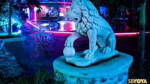 Una estatua de león de piedra en un jardín, con un edificio iluminado al fondo.