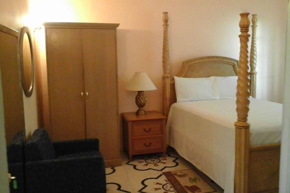 A bedroom with a four-poster bed, a wooden wardrobe, a bedside table with a lamp, and a chair.